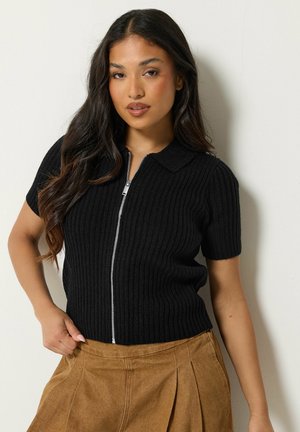 Femme aux longs cheveux foncés porte un haut noir côtelé à manches courtes avec fermeture éclair et un pantalon plissé marron, debout contre un mur clair.