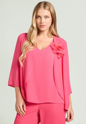 Blondynka w jasnoróżowej bluzce z dekoltem w serek i dopasowanych spodniach, z dużym kwiatem z tkaniny na lewym ramieniu.