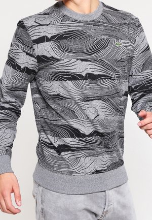 Harmaa paita, jossa on musta abstrakti puunsykuvio ja pieni vihreä krokotiililogo, päällä henkilö, jolla on vaaleanharmaat housut.