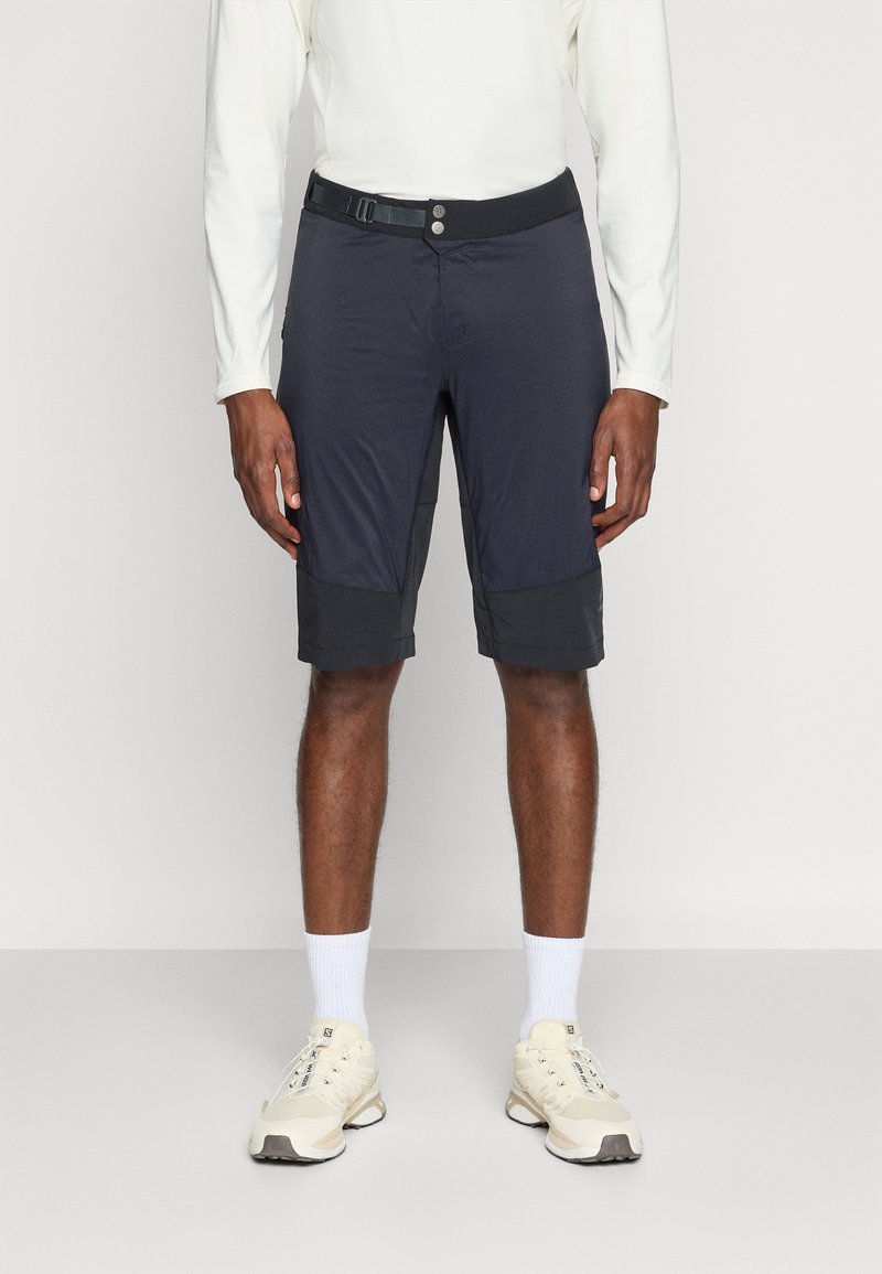 Ein Mann trägt schwarze knielange Shorts mit eingebautem Gürtel, ein weißes langärmliges Hemd, weiße Socken und beige Turnschuhe und steht vor einem einfarbigen Hintergrund.