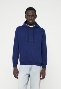 Hoodie bleu marine en tissu doux, avec une poche avant, un cordon de serrage ajustable et un logo "ORIGINALS" en relief. Porté avec un jean clair.