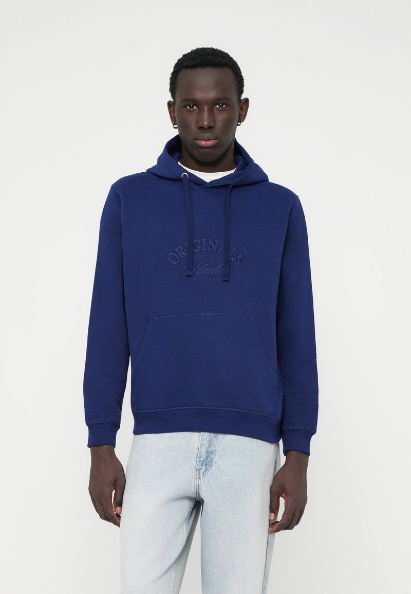 Hoodie bleu marine en tissu doux, avec une poche avant, un cordon de serrage ajustable et un logo "ORIGINALS" en relief. Porté avec un jean clair.
