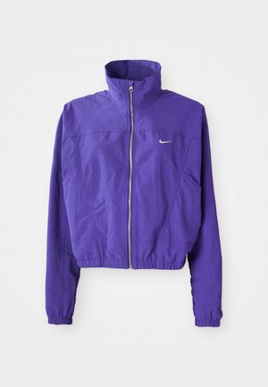 Lilla zip-up jakke laget av lett stoff, med høy krage og elastisk kant, med en liten logo på venstre bryst.