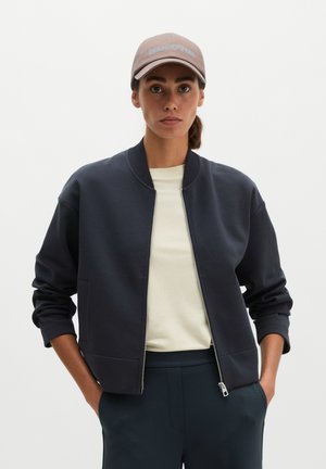 Femme portant une casquette beige, une chemise crème et une veste zippée bleu marine, les mains dans les poches, debout devant un fond clair uni.