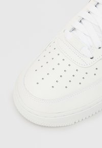 Fehér tornacipő perforált orr résszel, szintetikus anyagból, texturált talppal, lapos fűzőkkel és fém díszítéssel a szellőzés érdekében.