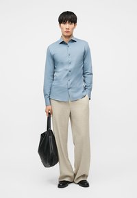 Muž v svetlomodrej košeli na gombíky a béžových širokých nohaviciach drží čiernu koženú tašku na rameno a stojí na bielom pozadí.