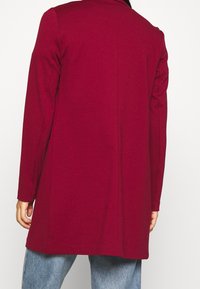 Manteau bordeaux à manches longues avec une coupe droite et un col cranté, présentant une texture lisse et des détails de couture minimalistes au dos.
