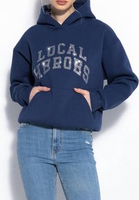 Granatowa, oversize bluza z kapturem z napisem "LOKALNY BOHATER" wykonanego z błyszczących srebrnych kryształków, z kieszenią z przodu i ściągaczami, zestawiona z jasnoniebieskimi dżinsami.