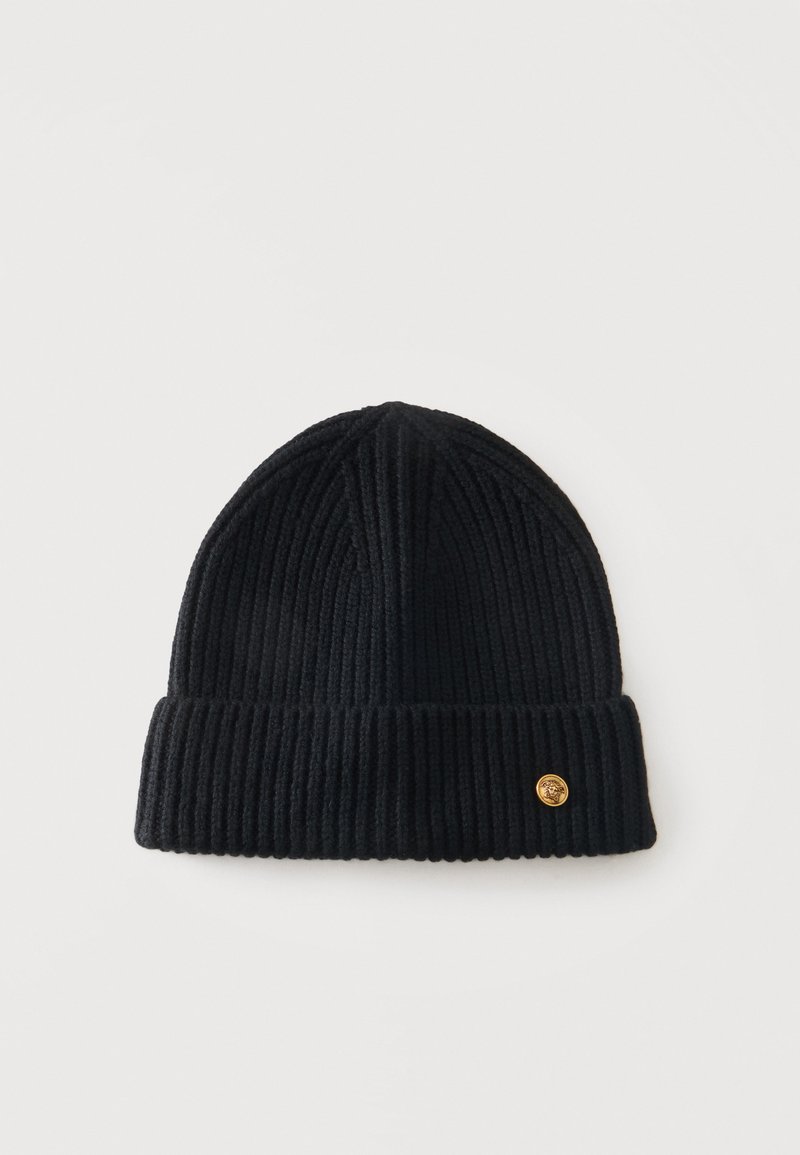 Bonnet en maille côtelée noire avec revers plié et petit emblème rond doré sur le revers sur fond blanc.
