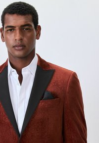 Roter strukturierten Blazer mit schwarzen Revers, kombiniert mit einem weißen Hemd. Das Einstecktuch hat ein einfarbig schwarzes Muster und verleiht einen formellen Look.