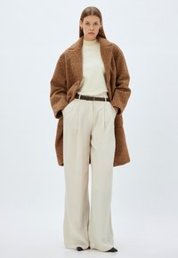 Brun uldfrakke over en cremefarvet sweater, højtaljede beige bukser og mørke stiletter. Har brede ben og en belagt talje.
