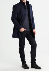 Marineblauer Wollmantel mit hohem Kragen und Knopfverschluss, kombiniert mit einem grauen Pullover, dunklen Jeans und schwarzen Lederstiefeln.