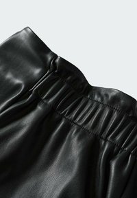 Vêtement en similicuir noir avec un détail de couture élastique froncée près du bord supérieur, présenté sur un fond clair uni.