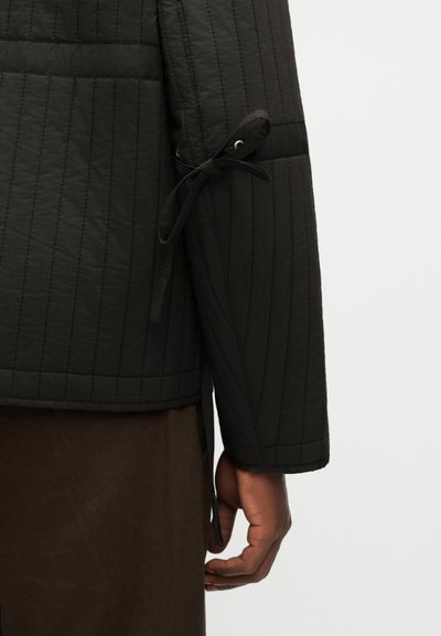 Manche de veste noire matelassée avec un détail de nœud au-dessus du poignet, portée sur une jupe ou un pantalon marron foncé, avec une main visible reposant sur le côté.