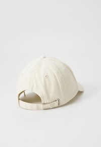 Beige bomullslue med buet brem, justerbar stropp bak, og sydd ventilasjonsåpninger. Teksturert stoff med et enkelt design.