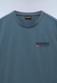 Camiseta de algodón en color teal con cuello redondo. Presenta un logo impreso, texto y un pequeño gráfico de bandera en el pecho. Textura suave.