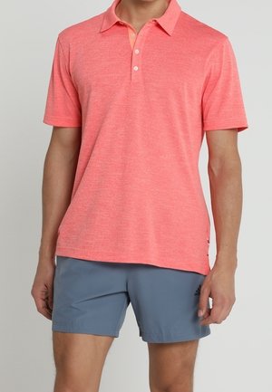 Man wearing a coral pink short-sleeve polo shirt and gray Adidas shorts, standing against a plain light background.