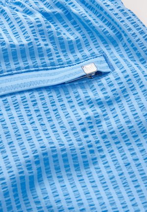 Lyseblåt stof med lodrette hvide striber, der har en lynlåslomme og en tekstureret overflade. Sølvfarvet lynlåshardware tilføjer detaljer.