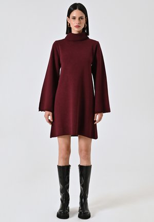 Abito a maglia color marrone scuro con collo alto, maniche a campana lunghe e orlo sopra il ginocchio, abbinato a stivali neri fino al ginocchio.