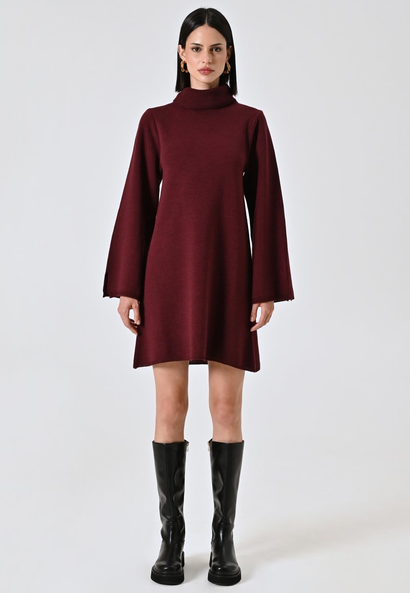 Abito a maglia color marrone scuro con collo alto, maniche a campana lunghe e orlo sopra il ginocchio, abbinato a stivali neri fino al ginocchio.