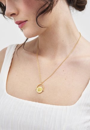 Kobieta o ciemnych włosach w białej bluzce nosi złoty naszyjnik z okrągłą zawieszką przedstawiającą wzór rozchodzącego się promieniowania słonecznego.