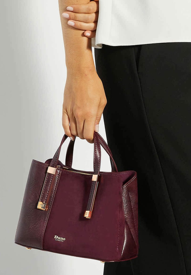 Bordeaux neplederen handtas met twee korte handvatten, gouden hardwareaccenten en een ritsluiting. Textuur body met gladde panelen.