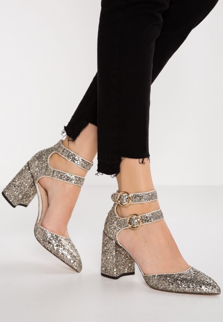 Chaussures à talons hauts argentées étincelantes avec doubles brides à la cheville et boucles dorées, portées avec un pantalon noir à ourlet effiloché sur un fond uni.