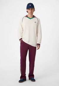 Kermainen pusero, jossa on viininpunaisia ja vihreitä yksityiskohtia, yhdistettynä viininpunaisiin housuihin, joissa siniset sivuraidat. Asusteena tummansininen lippalakki.