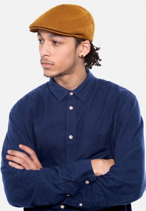 Uomo con coppola marrone chiaro, camicia blu con bottoni, braccia incrociate, guarda a sinistra su sfondo bianco uniforme.