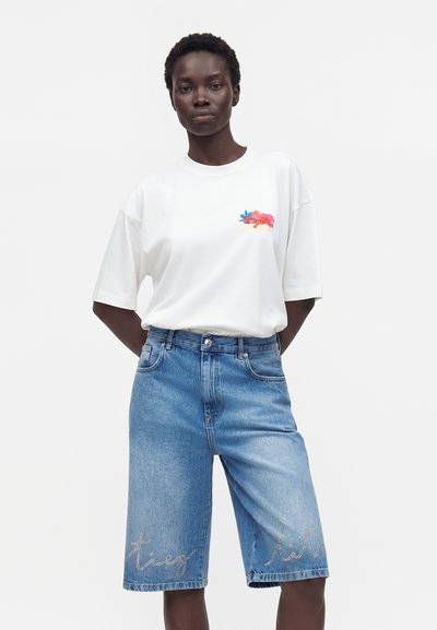 Γυναίκα φοράει λευκό oversized T-shirt με μικρό πολύχρωμο λουλουδάτο λογότυπο και ψηλόμεσα μπλε denim σορτς μήκους μέχρι το γόνατο με κείμενο από στρας.