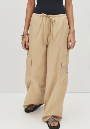 Pantaloni cargo larghi beige con elastico in vita e cordino, dotati di due tasche laterali e un'etichetta in tessuto sulla coscia sinistra.