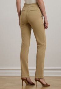 Γυναίκα με μπεζ στενά παντελόνια και καφέ ψηλοτάκουνα σανδάλια, standing σε ξύλινο δάπεδο μπροστά σε έναν ανοιχτό τοίχο.