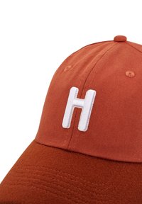 Rostfärgad keps i bomullsmaterial, med en texturerad, mjuk tyg och en framträdande vit broderad "H" på framsidan.