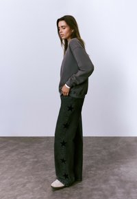 Mujer de pie de lado, vistiendo un sudadera gris holgada, pantalones vaqueros negros con estampados de estrellas negras en la pierna y zapatillas blancas.