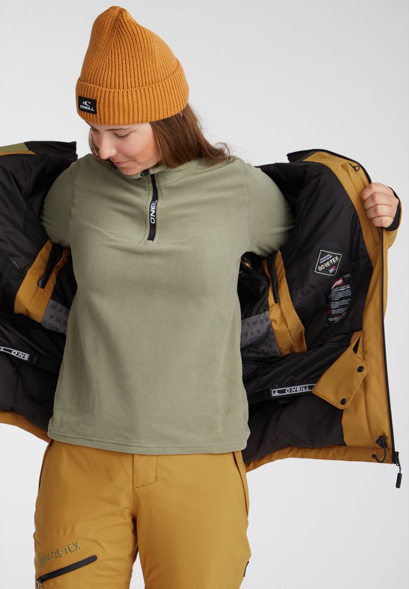 Olivgrüner Fleece-Pullover mit einem halben Reißverschluss, kombiniert mit gelben Hosen und einer gerippten, senfgelben Mütze, unter einer schwarz-goldenen Kapuzenjacke.