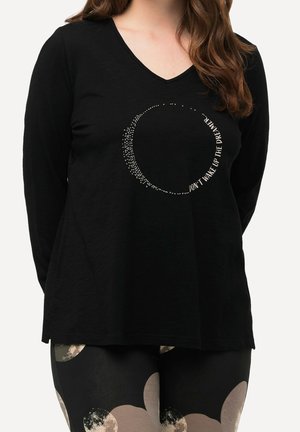 Kobieta ubrana w czarną bluzkę z długim rękawem i dekoltem w serek, ozdobioną kolistym wzorem z kropek oraz napisem „Nie budź marzycielki.” Czarne legginsy z beżowymi wzorami.