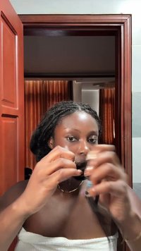 Person med mörkt, lockigt hår som håller en flaska och bär en vit handduk. I bakgrunden syns en trädörr och bruna gardiner.