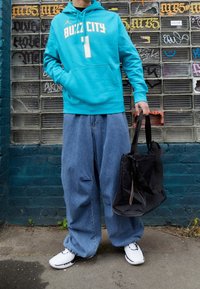 Sweat à capuche turquoise avec lettrage blanc, pantalon en denim bleu ample, et sac noir. Chaussures blanches avec des accents noirs.