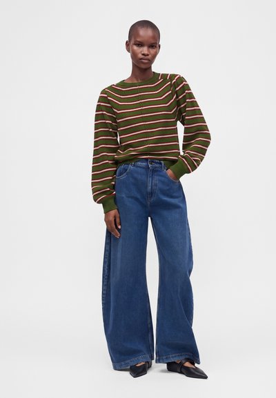Modèle portant un pull rayé vert et un jean bleu à jambes larges, une main dans la poche, debout sur un fond blanc uni.