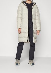 Ljusgrå lång pufferjacka med huva, med horisontell kviltad söm, frontdragkedjestängning och elastiska ärmmuddar. Bar över mörka byxor.