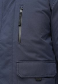 Granatowa ocieplana kurtka z suwakiem na kieszeni na piersi, płaską kieszenią z przodu oraz fakturą tkaniny z detalami przeszyć.