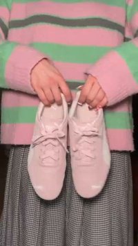 Helle rosa Sportschuhe aus glattem Material, mit Schnürung und gepolsterter Sohle, gehalten von Händen vor einem gestreiften Pullover.