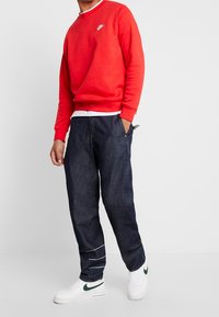 Röd crewneck-tröja med en vit Nike-logga, kombinerad med lätt lösa mörk denimbyxor och vita sneakers med gröna accenter.