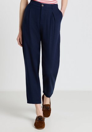 Frau trägt marineblaue plissierte, zulaufende Hosen mit Seitentaschen und braune Wildleder-Schnürslipper, steht vor einem schlichten hellen Hintergrund.