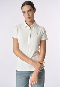 Polo bianco con colletto, maniche corte e patta con tre bottoni. Presenta un piccolo logo ricamato sul petto. Tessuto liscio.