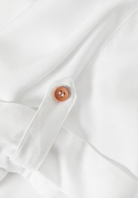 Tela blanca con una textura suave, que presenta un botón decorativo marrón con dos agujeros, colocado en una sección doblada.