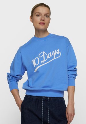 Kvinde iført en blå sweatshirt med hvid tekst ""10 Days"", parret med mørke bukser med snøre, stående med den ene hånd i lommen.