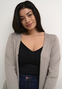 Cardigan beige sopra una maglietta nera attillata con un design arricciato, indossato con jeans in denim scuro. Texture liscia, design casual e semplice.
