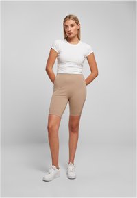 Beige fiets-short met een elastische, gladde textuur, gecombineerd met een aansluitend wit T-shirt. Model draagt witte sneakers. Eenvoudig ontwerp.