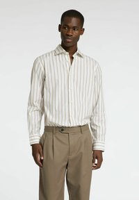 Biała koszula z guzikami, z pionowymi paskami w odcieniach beżu i jasnobrązowym, zestawiona z jasnobrązowymi spodniami. Koszula ma spiczasty kołnierzyk oraz wywinięte rękawy.